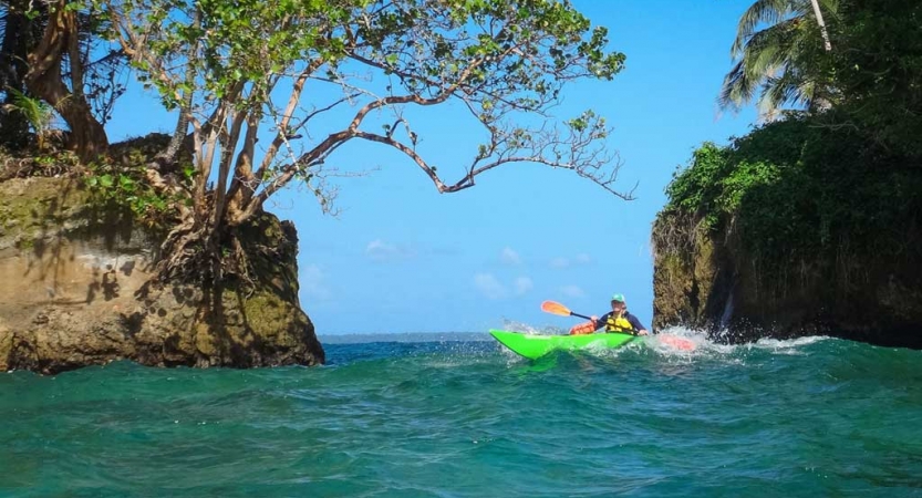 A person paddles a kayak through blue water between two tall rocks covered with trees and greenery.